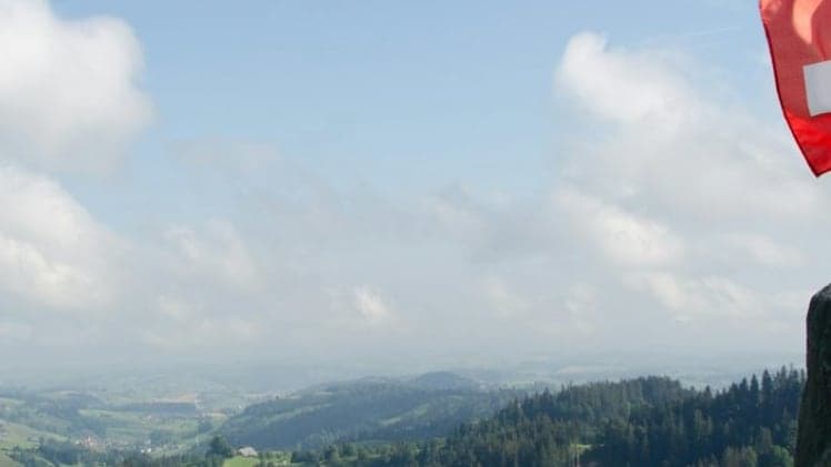 Swiss flag above green hills and forest under a cloudy sky