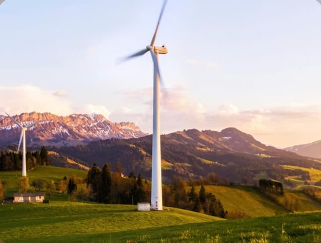 Wind turbines on green hills in front of a mountain range at sunset